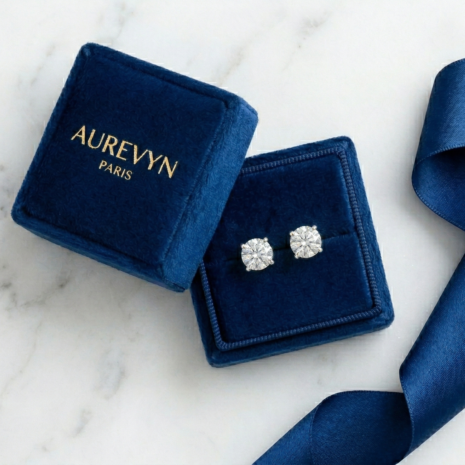 Diamond earrings in a blue Aurevyn Paris box on a marble surface with a blue ribbon.